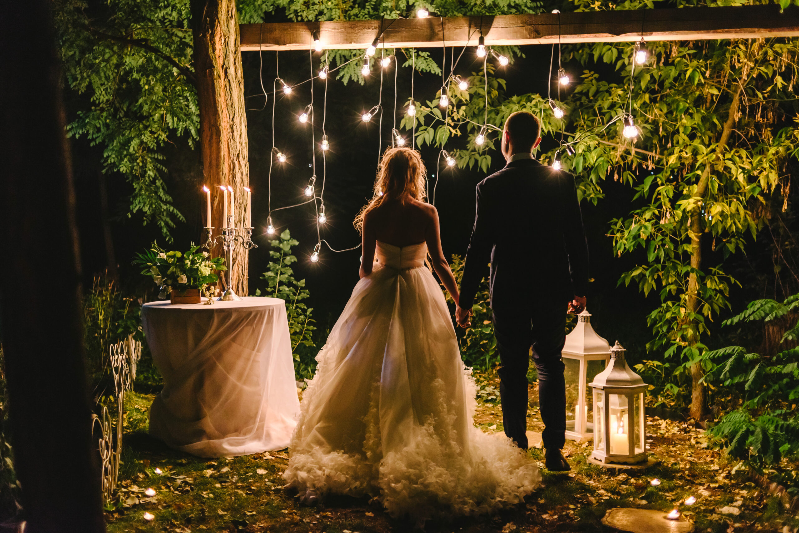 Night wedding ceremony with candles, lanterns and lamps on tree. Bride and groom holding hands on background of baulb lights, back view. Beautiful young couple standing under a tree at night